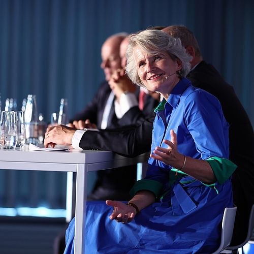 Fünf Personen sitzen bei einer Konferenz zusammen und eine Frau spricht in Richtung der Kamera.