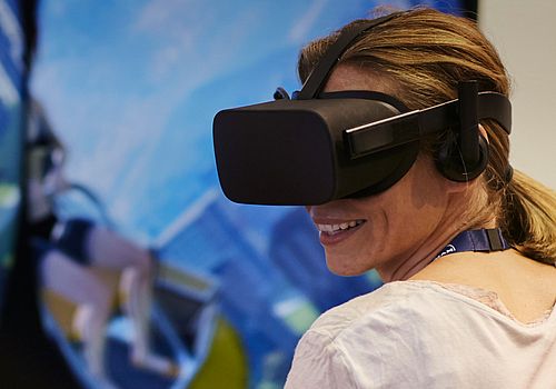 Eine lächelnde Frau, die ein VR-Headset trägt. Verlinkt zu "Themenrouten"