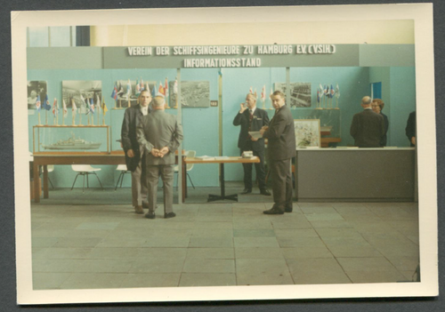 Historisches Bild des Informationsstandes des Vereins der Schiffsingenieure zu Hamburg E.V.. Mit zwei Tischen und mehreren Bildern an der Wand hängend.