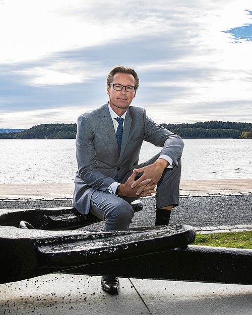 Ein Mann in grauem Anzug sitzt auf einem großen Anker. Hinter ihm ist eine Landschaft mit einem Gehweg, Wasser, einem blau bewölktem Himmel und grünem Festland zu sehen.