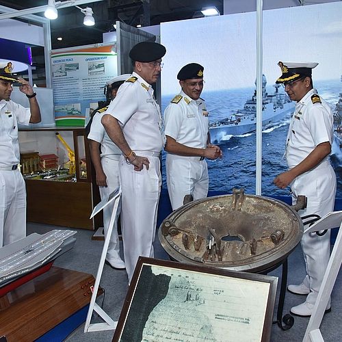 Das Bild zeigt eine Gruppe von Marineoffizieren in Uniform, die sich auf einer Ausstellung über maritime Technologie unterhalten. Im Hintergrund ist ein großes Bild von Kriegsschiffen auf See zu sehen, und es werden Modelle von Schiffen sowie historische maritime Artefakte ausgestellt.