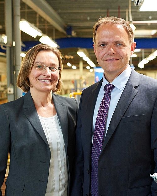 Tanja und Ronald Hoppmann schauen lächelnd in die Kamera und tragen graue Anzugsjacken. Im Hintergrund lässt sich eine Art Lagerhalle erahnen.